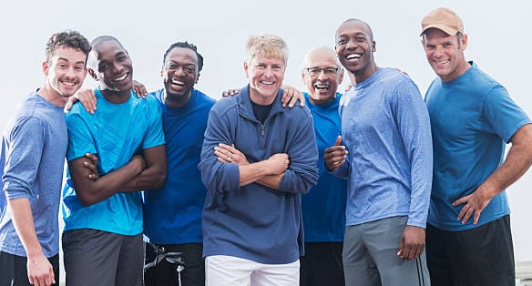 Diverse group of men wearing casual blue shirts, standing together outdoors.  Mixed ages, 20s to 70s.  They are confident and happy, smiling at the camera.