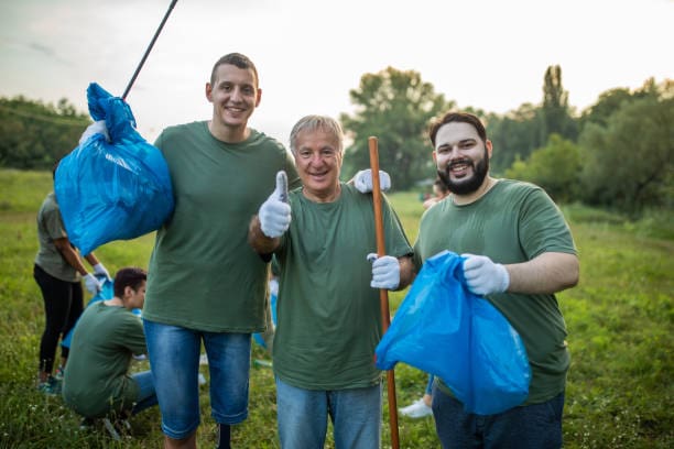 Portrait of three male volunteers, standing, embracing, smiling and looking at camera, after cleaning public park, they have same green t-shirts,